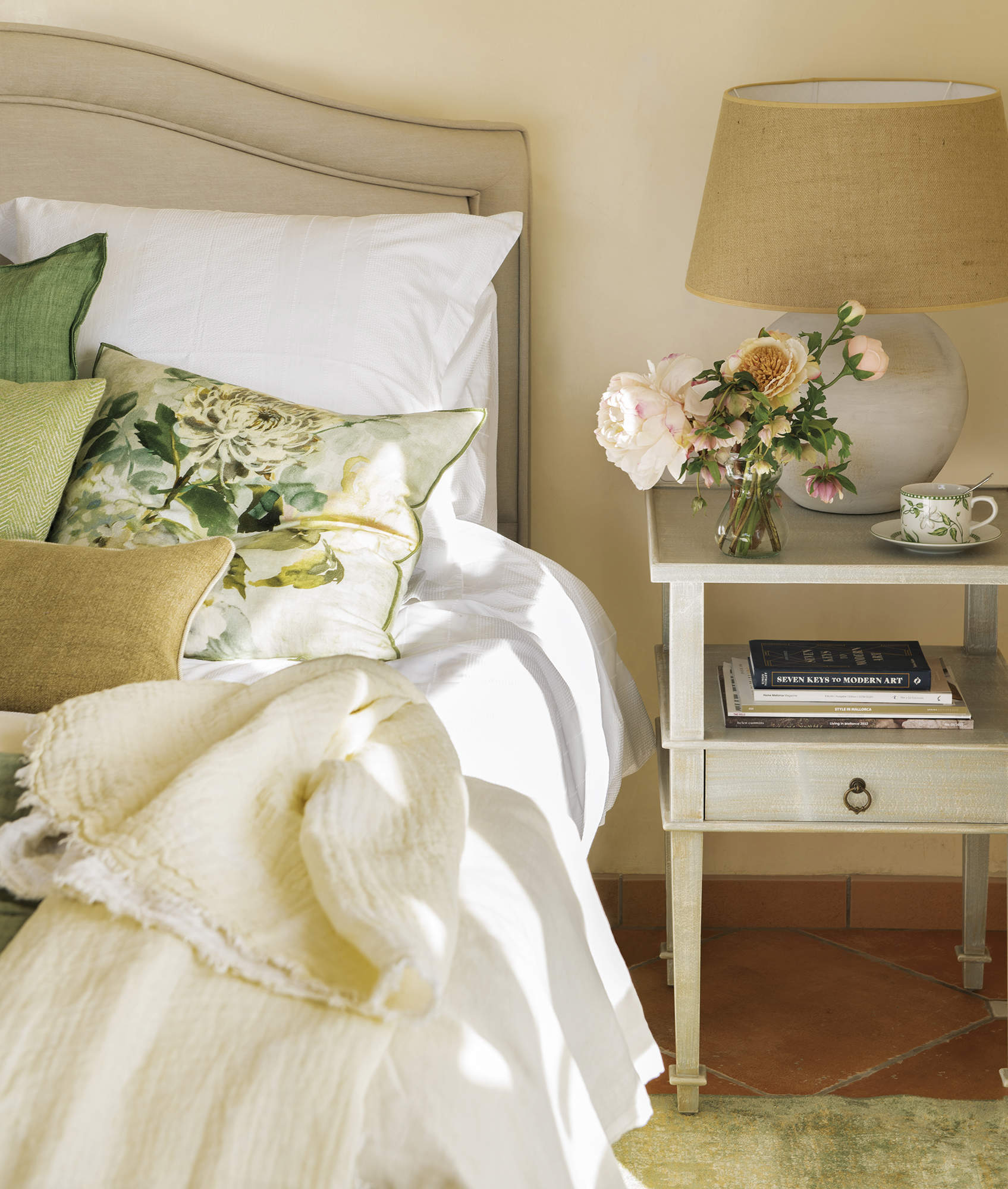 dormitorio con cabezal tapizado, mesa de noche con lámpara y flores, ropa de cama blanca y verde con flores